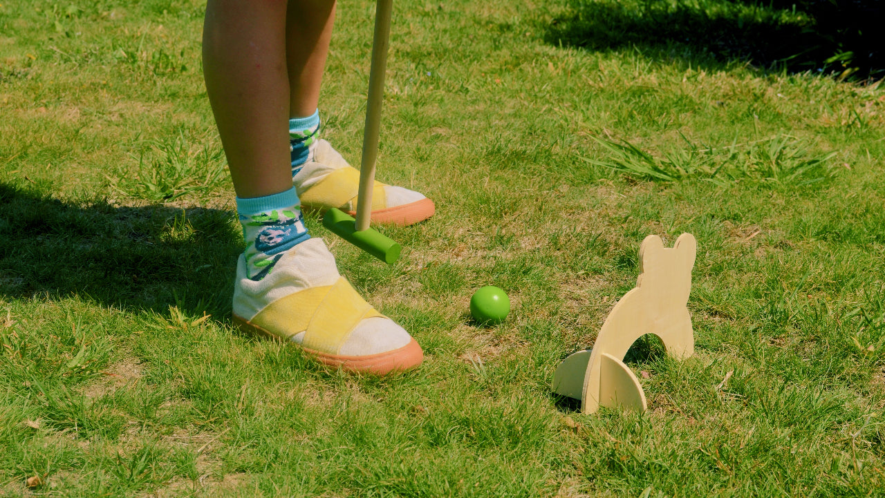 child playing croquet on a grassy lawn with a mallet and ball, wearing yellow canvas sustainable shoes