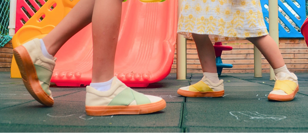 Children's feet wearing colorful sustainable kids shoes on a playground with slides and play equipment in the background.