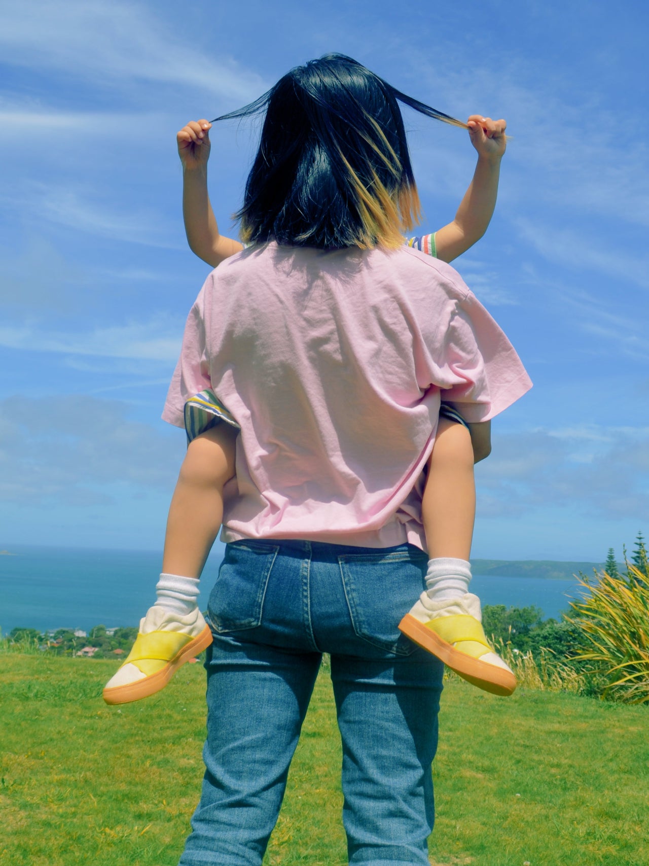 Mother holding a child on their shoulders with a clear blue sky and ocean in the background. Child wears yellow canvas sustainable shoes
