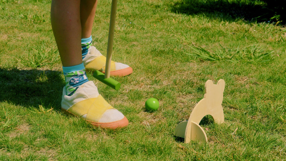 Kid wearing sustainable sneakers playing croquet on a grassy lawn with a mallet and ball.