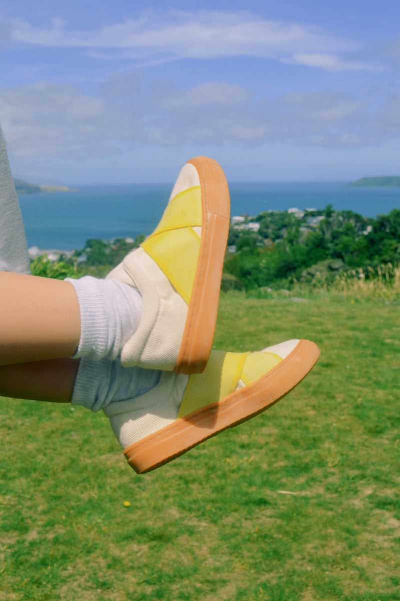 Kid wearing yellow sandals with brown soles on a grassy field with a scenic background.