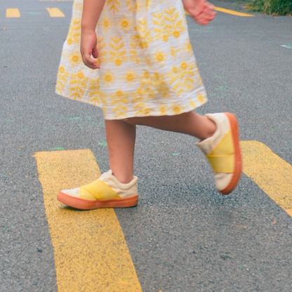 Child wearing a white dress with yellow floral patterns and matching sustainable kids shoes walking on a crossing road.